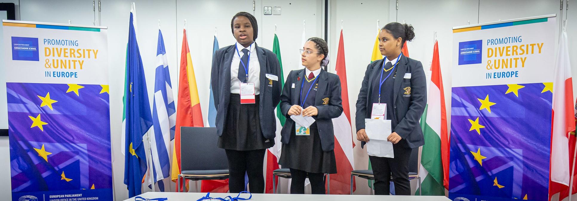 3 Loreto students presenting in front of EU flags